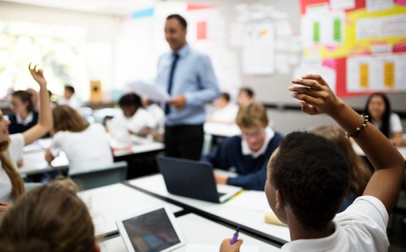 male teacher standing in clasroom students raising hands