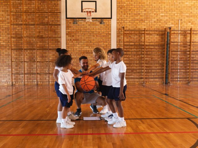 Schoolkids and basketball coach forming hand stack at basketball court in school